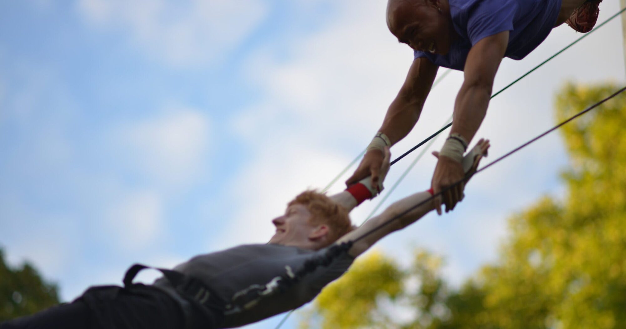 One man on a swinging bar holds the wrists of another man who is swinging in the air